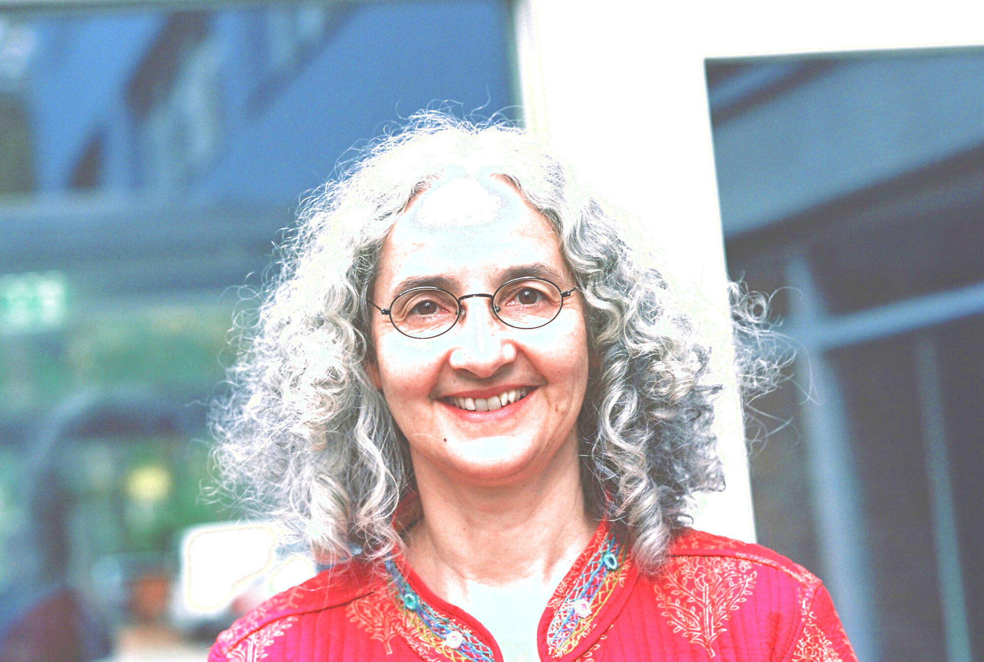 Das Porträt zeigt eine lachende Frau (Carola Bauckholt). Sie schaut geradeaus und trägt ein rotes Kleid.