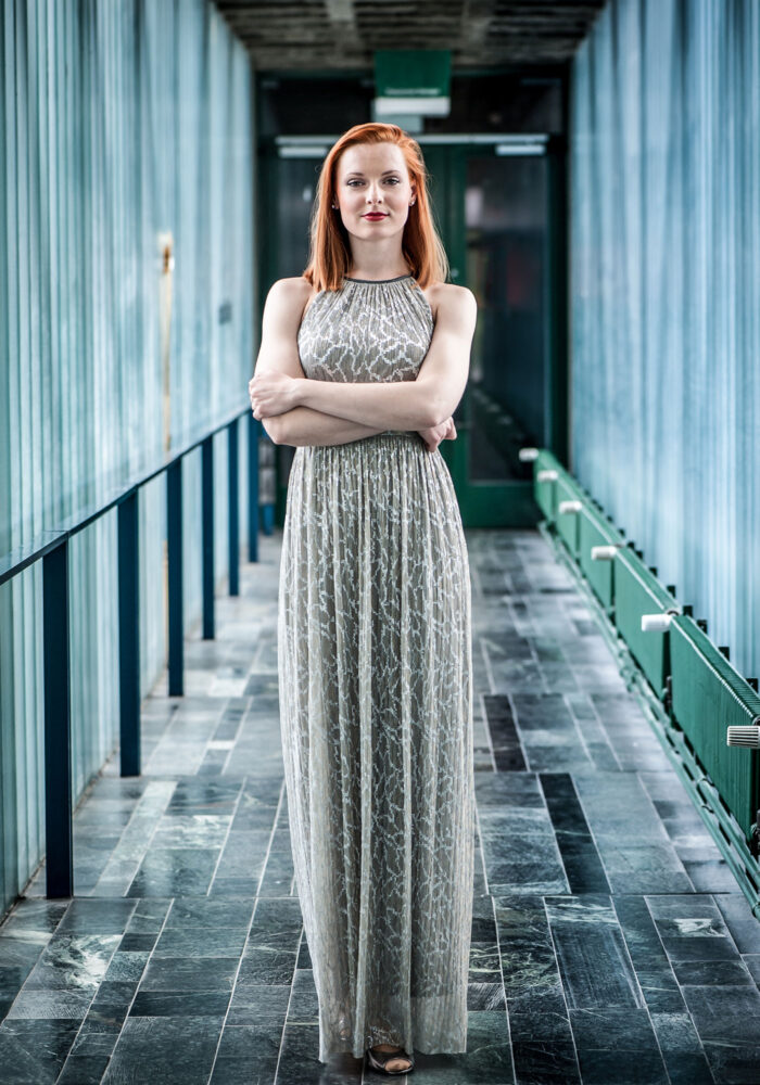 Das Bild zeigt eine Frau in einem grauen, paillettenbesetzten Abendkleid (Viktoriia Vitrenko). Ihre Arme sind in der Taille verschränkt. Sie steht in einem Gang aus Glas und Metall.