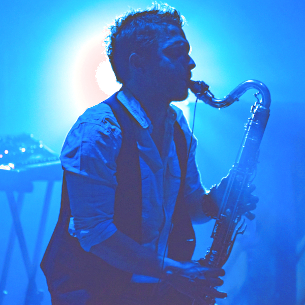 Das Bild zeigt einen Mann, den Musiker Gareth Davis. Er steht seitlich und spielt Saxophon. Das Bild wurde während seines Auftritts auf der Bühne aufgenommen. Er trägt ein blaues Hemd und eine schwarze Weste.
