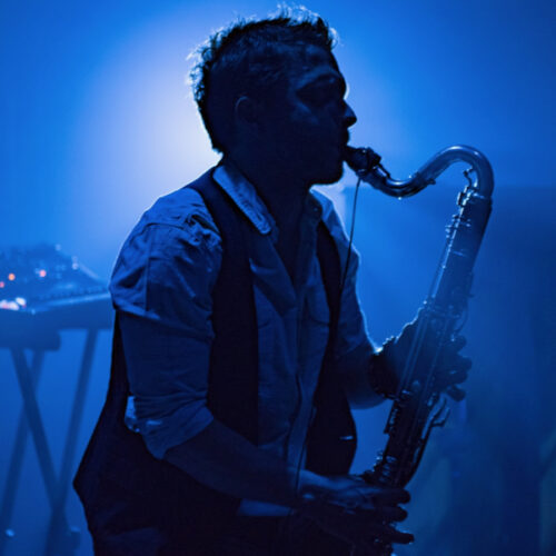 Das Bild zeigt einen Mann, den Musiker Gareth Davis. Er steht seitlich und spielt Saxophon. Das Bild wurde während seines Auftritts auf der Bühne aufgenommen. Er trägt ein blaues Hemd und eine schwarze Weste.