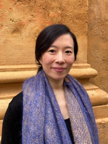 Zu sehen ist ein Portrait der Komponistin Huihui Cheng. Sie steht vor einer Säule aus Sandstein und trägt ein schwarzes Oberteil und einen lilanen Schal.
