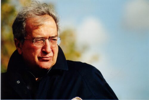 Zu sehen ist ein Portrait des Komponisten Luciano Berio. Im Hintergrund sind verschwommen Bäume und der blaue Himmel zu sehen. Er trägt einen dunklen Mantel und blickt zur Seite.