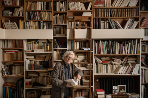Zu sehen ist der Komponist George Aperghis. Mit einem Buch in der Hand lehnt er an einer Leiter vor einer Wand voll mit Büchern.
