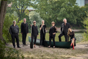 An einem Flussufer stehen die sieben Sänger:innen der Neuen Vocalsolisten um ein grünes Kanu herum. Zwei von ihnen halten je ein Paddel. Sie tragen alle schwarze Konzertkleidung.
