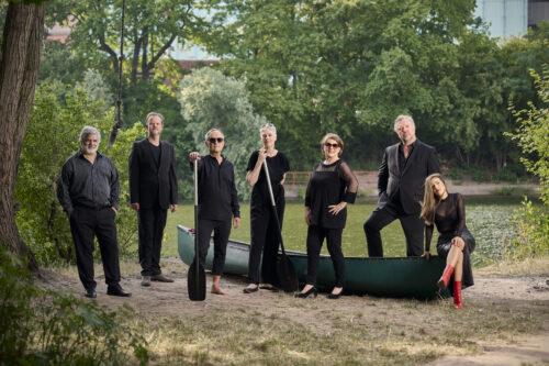 An einem Flussufer stehen die sieben Sänger:innen der Neuen Vocalsolisten um ein grünes Kanu herum. Zwei von ihnen halten je ein Paddel. Sie tragen alle schwarze Konzertkleidung.