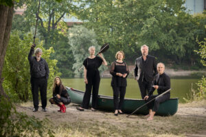 An einem Flussufer stehen sechs Sänger:innen der Neuen Vocalsolisten um ein grünes Kanu herum. Sie tragen alle schwarze Konzertkleidung.