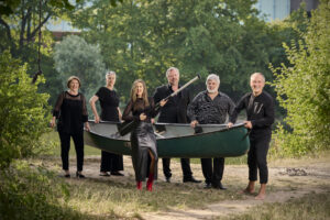 An einem Flussufer stehen sechs Sänger:innen der Neuen Vocalsolisten. Sie halten gemeinsam ein grünes Kanu. Die Frau im Vordergrund hält ein Paddel. Sie tragen alle schwarze Konzertkleidung.