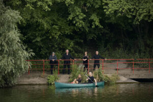 Zu sehen sind sechs Sänger:innen der Neuen Vocalsolisten. Zwei von ihnen sitzen in einem grünen Kanu auf dem Fluss. Die restlichen vier stehen nebeneinander am Flussufer direkt hinter dem Kanu.