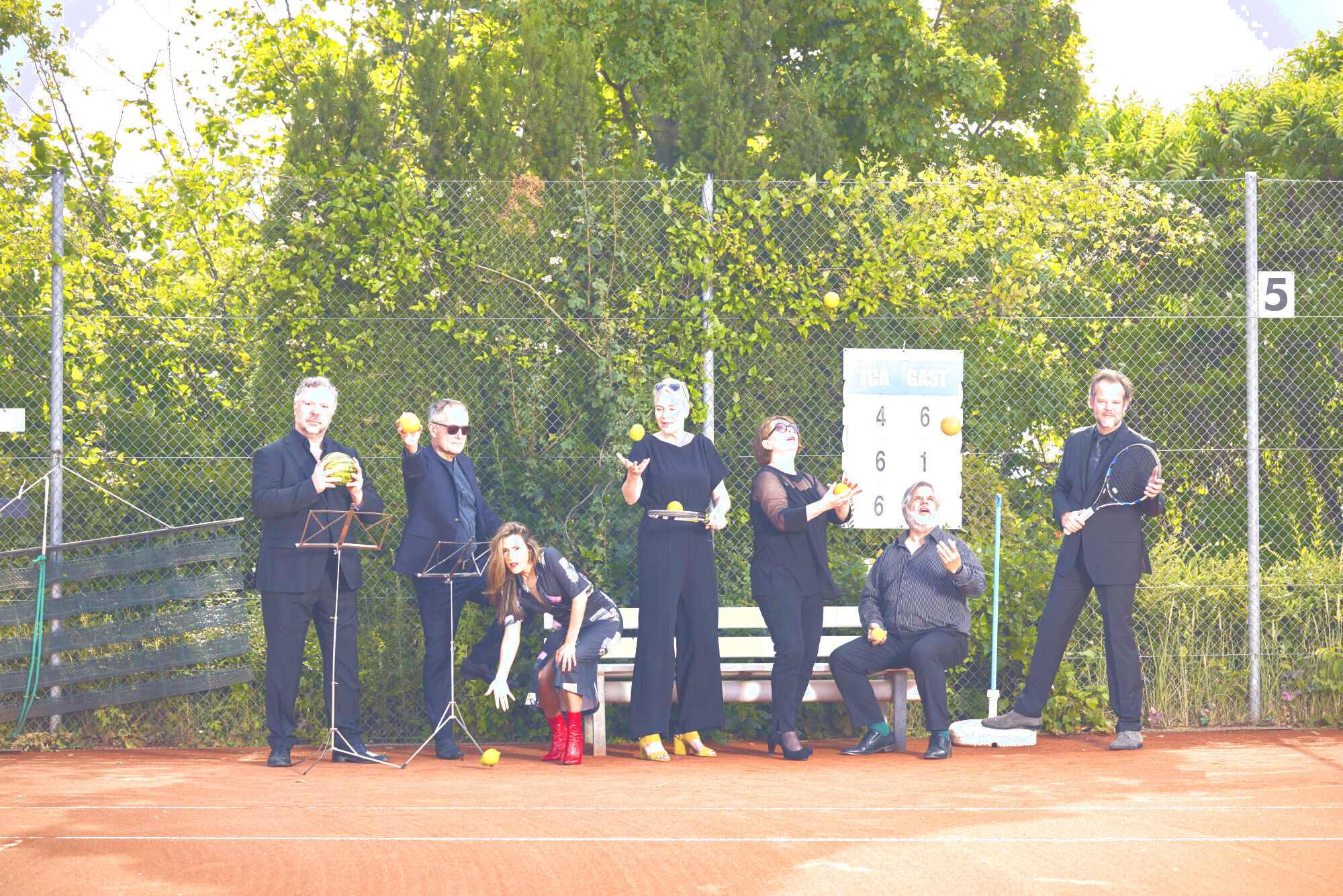 Auf dem Bild steht die Gruppe von sechs Sänger:innen der Neuen Vocalsolisten. Sie stehen auf dem Tennisplatz und halten Orangen, Zitronen, Äpfel, Melonen und Tennisschläger in den Händen.