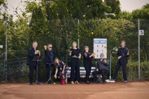 Auf dem Bild steht die Gruppe von sechs Sänger:innen der Neuen Vocalsolisten. Sie stehen auf dem Tennisplatz und halten Orangen, Zitronen, Äpfel, Melonen und Tennisschläger in den Händen.