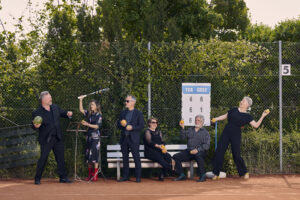 Auf dem Bild steht die sechs Sänger:innen der Neuen Vocalsolisten. Sie tragen Konezrtkleidung und. stehen auf einem Tennisplatz. Sie halten Orangen, Zitronen, Äpfel, Melonen und Tennisschläger in den Händen.