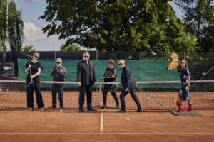Die sechs Sanger:innen der Neuen Vocalsolisten befinden sich auf einem Tennisplatz. Sie tragen Konzertkleidung. Ein Mann zieht eine Frau auf dem Schleppnetz. Sie hält einen Fächer. Die anderen halten Tennisbälle und Schläger sowie Notenständer in ihren Händen.