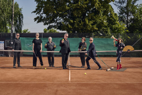 Die sechs Sanger*innen der Neuen Vocalsolisten befinden sich auf einem Tennisplatz. Sie tragen schwarze Kleidung. Ein Mann zieht eine Frau auf dem Schleppnetz. Sie hält ein Megaphon und einen Fächer. Die anderen halten Tennisbälle und Schläger sowie Notenständer in ihren Händen.