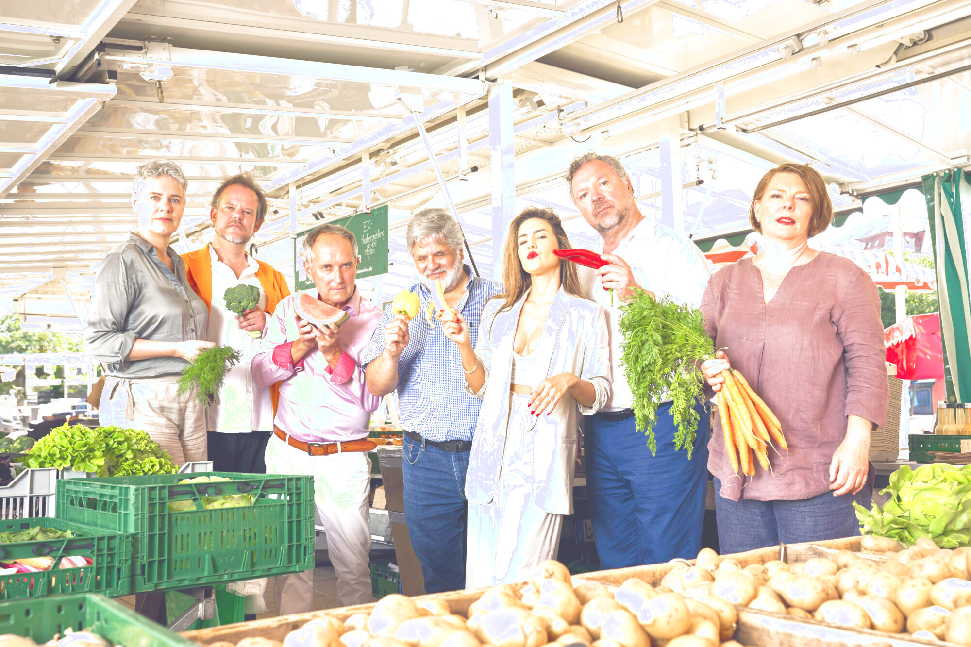 Zu sehen sind die sieben Sänger:innen der Neuen Vocalsolisten. Sie stehen inmitten eines Marktstandes. Jede:r Sänger:in hält ein Stück Obst oder Gemüse in der Hand.