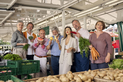 Zu sehen sind die sieben Neue Vocalsolisten. Sie stehen inmitten eines Marktstandes. Jede*r Sänger*in hält ein Stück Obst oder Gemüse in der Hand.