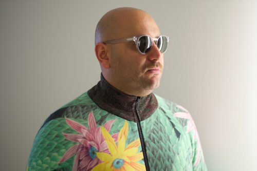 Zu sehen ist ein Portrait des Komponisten Raed Yassin. Er steht leicht schräg zur Kamera. Er trägt eine Sonnenbrille und eine grüne Jacke mit Pflanzenprint.