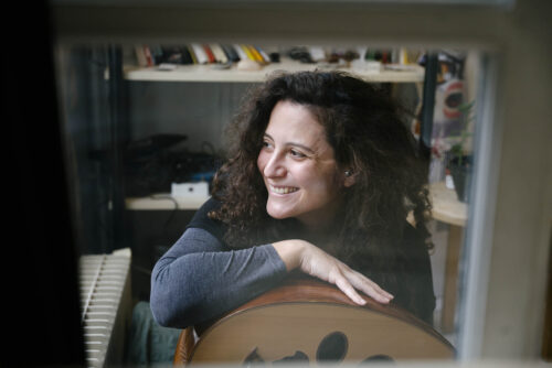 Zu sehen ist ein Portrait der Komponistin Youmna Saba. Sie sitzt an einem Klavier und hat auf ihrem Schoß eine Oud liegen. Sie hat ein breites Lächeln im Gesicht.