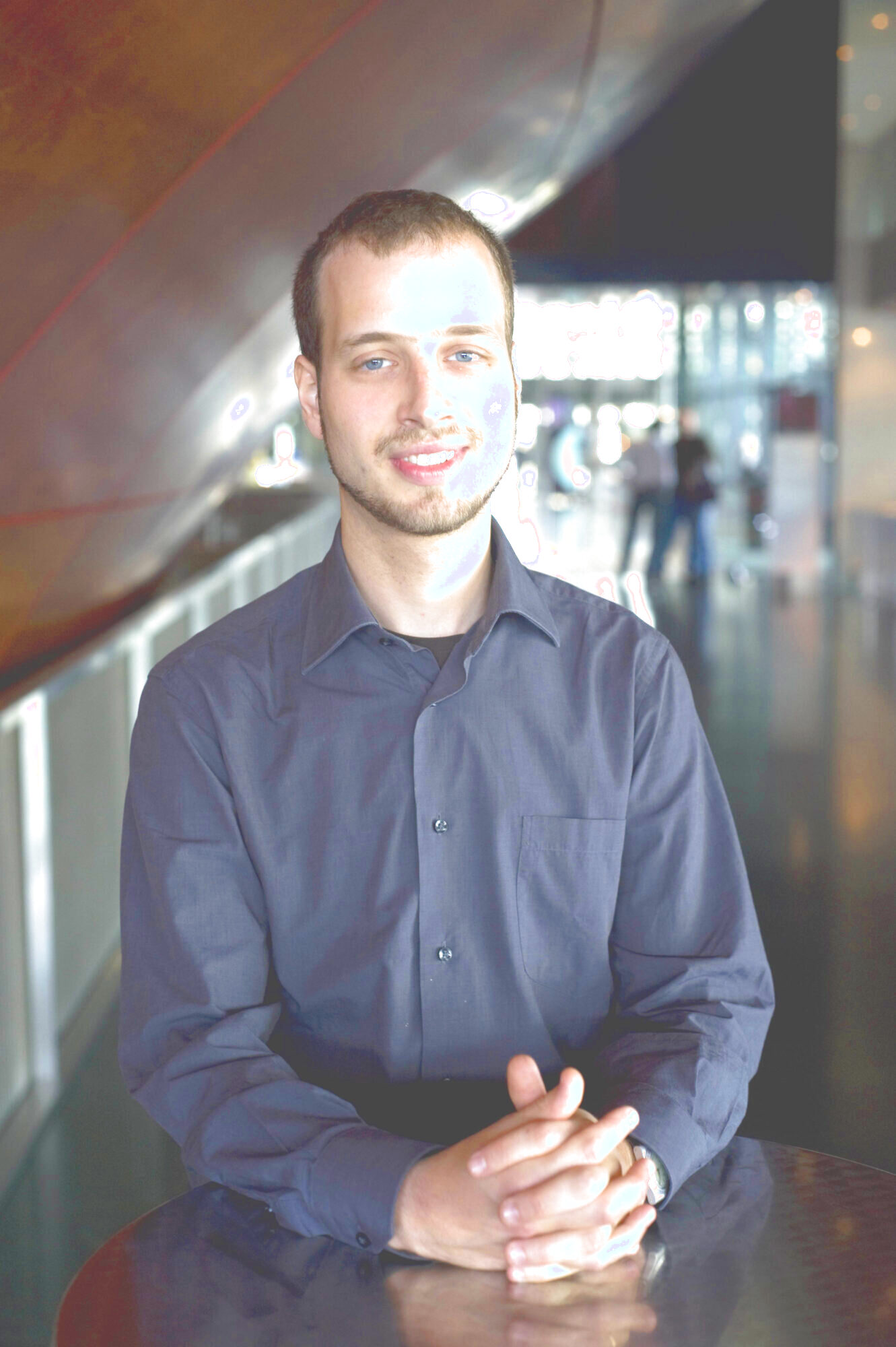 Das Porträt eines Mannes (Ondřej Adámek). Er sitzt an seinem Schreibtisch, die Hände vor sich gefaltet. Er blickt geradeaus und trägt ein schwarzes Hemd. Im Hintergrund ist das Innere eines öffentlichen Gebäudes zu erkennen.