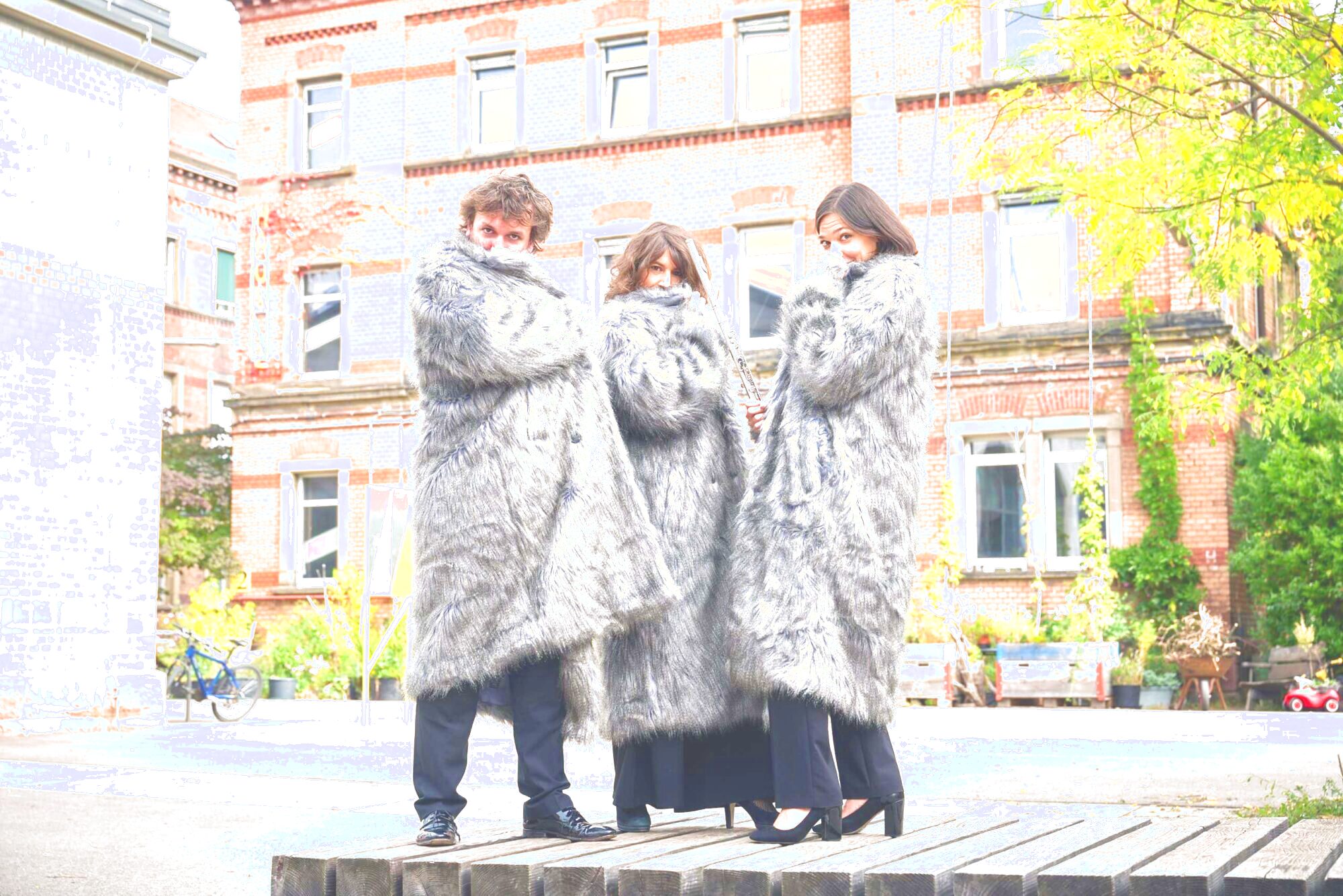 Das Bild zeigt zwei Frauen und einen Mann (TRIO vis-à-vis). Sie tragen Fellmäntel und verdecken damit ihre Gesichter. Im Hintergrund ist ein Gebäude zu erkennen.