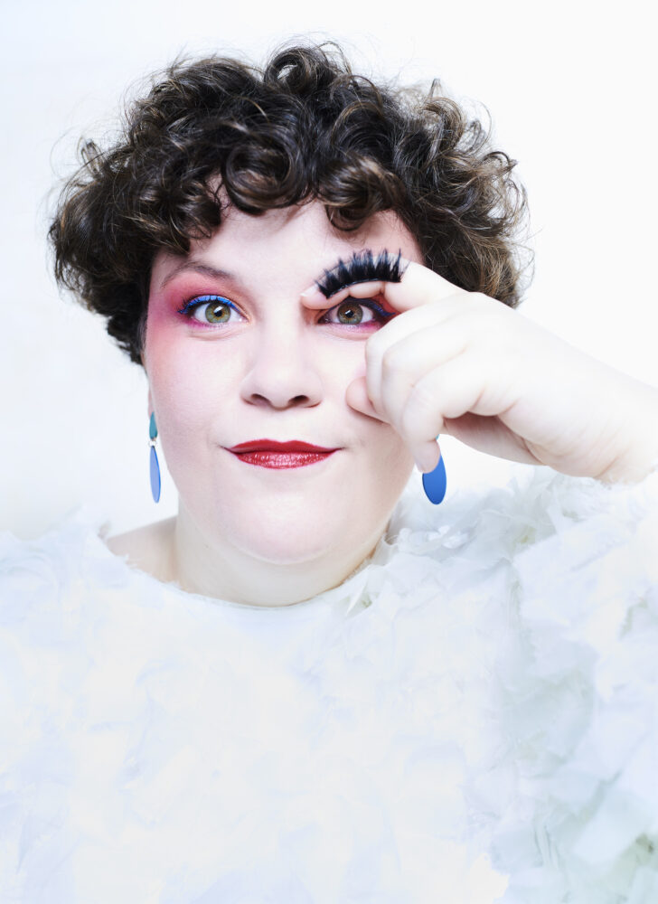 Porträt einer Frau im hellen Abendkleid (Andrea Conangla). Der Zeigefinger einer Hand ist gebogen und liegt wie eine Augenbraue über dem Auge. Darauf sind schwarze künstliche Wimpern geklebt.