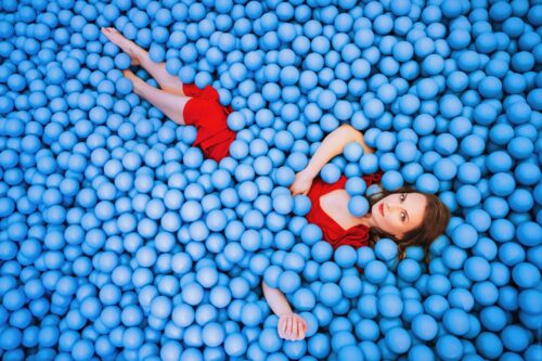 Das Bild zeigt eine junge Frau. Es ist die Sängerin Viktoriia Vitrenko. Sie liegt in einem Bällebad mit blauen Bällen. Sie trägt ein rotes Abendkleid.
