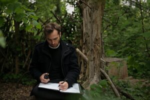 Der Komponist Arne Gieshoff. Er sitzt schreibend in einem Wald unter einem Baum.