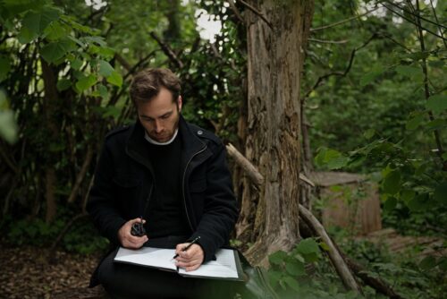 Der Komponist Arne Gieshoff. Er sitzt schreibend in einem Wald unter einem Baum.