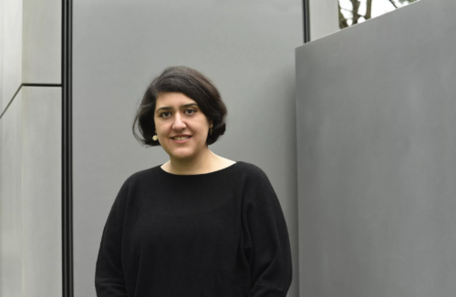 Zu sehen ist die Komponistin Elnaz Seyedi. Sie steht vor einer grauen Betonwand. Sie trägt ein schwarzes Oberteil und blickt lächelnd in die Kamera.