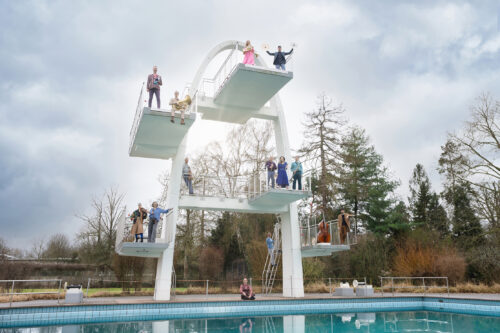 Zu sehen ist das Ensemble Musikfabrik. Verteilt sind die 13 Musiker*innnen mitsamt ihren Instrumenten auf den Sprungbrettern eines Sprungturmes in einem Freibad.