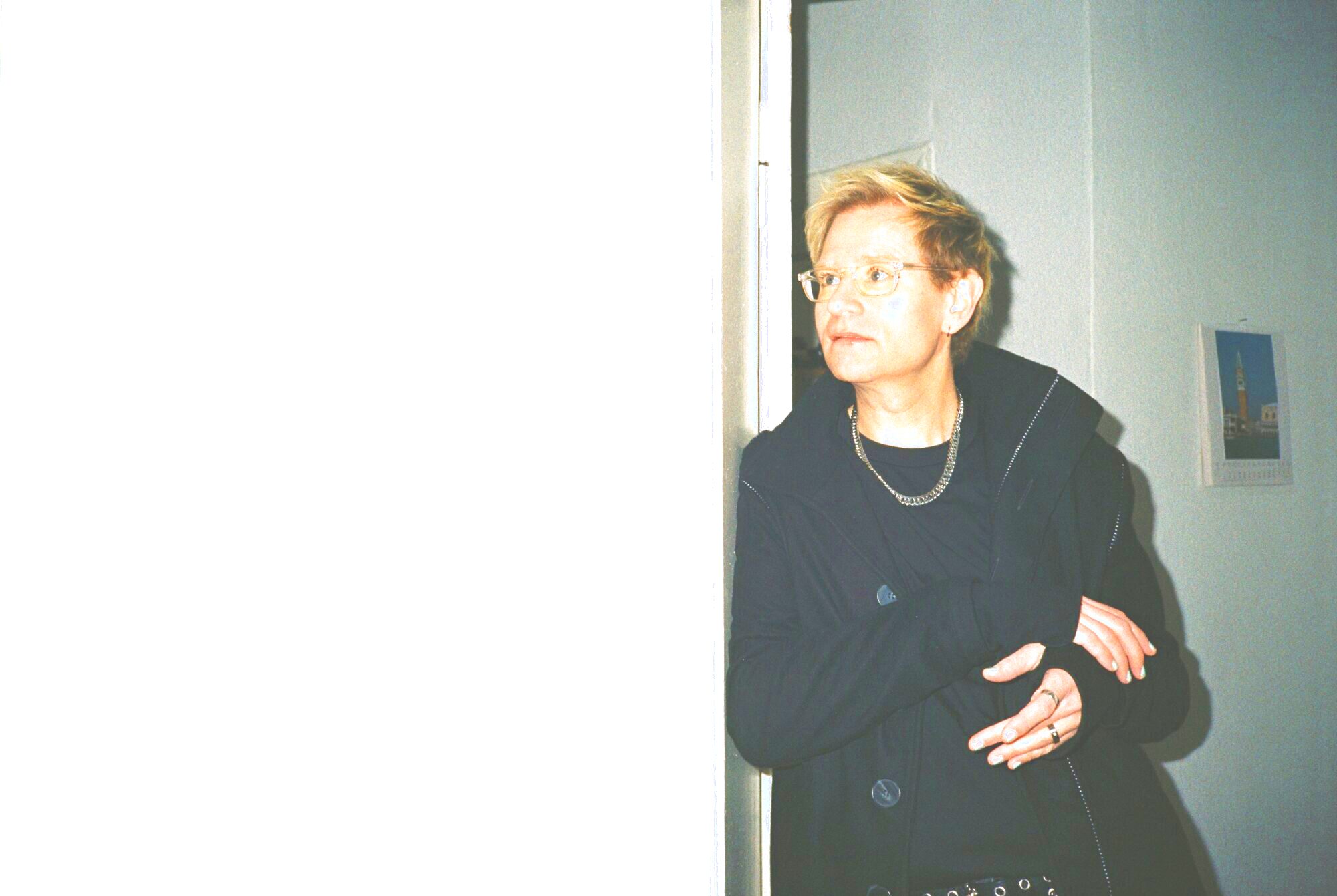 Das Bild zeigt Komponist*in Luxa Mart*in Schüttler. Luxa trägt einen schwarzen Mantel und lehnt an einer weißen Wand.