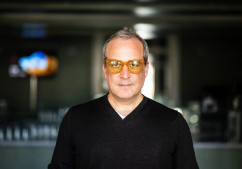 Zu sehen ist der Komponist Marc Sinan. Er trägt ein schwarzes Oberteil und eine orangefarbene getönte Brille.