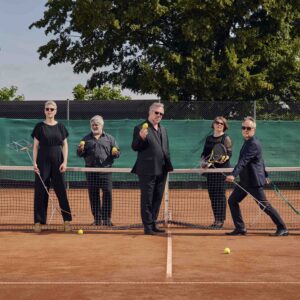 Das Bild zeigt fünf Sänger:innen der Neue Vocalsolisten auf einem roten Tennisplatz, alle in schwarzer Konzertkleidung. Zwei von ihnen halten Zitronen, zwei halten Notenständer und eine einen Tennisschläger. Eine Person zieht ein Schleppnetz.