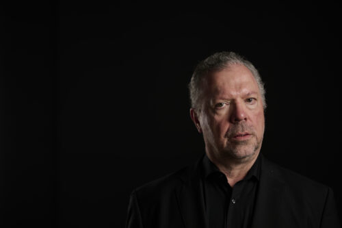 Das Bild zeigt ein Porträt von Andreas Fischer. Er steht vor einem schwarzen Hintergrund und trägt ein schwarzes Oberteil. Er blickt in die Kamera.