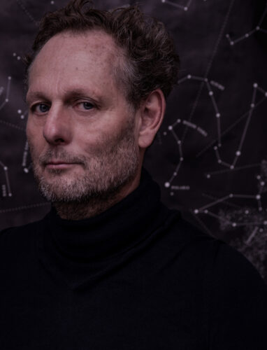 Zu sehen ist ein Portrait von Olaf NIcolai. Er steht vor einem dunklen Hintergrund. Er trägt ein schwarzes Oberteil. Im Hintergrund ist eine Sternkarte zu sehen.