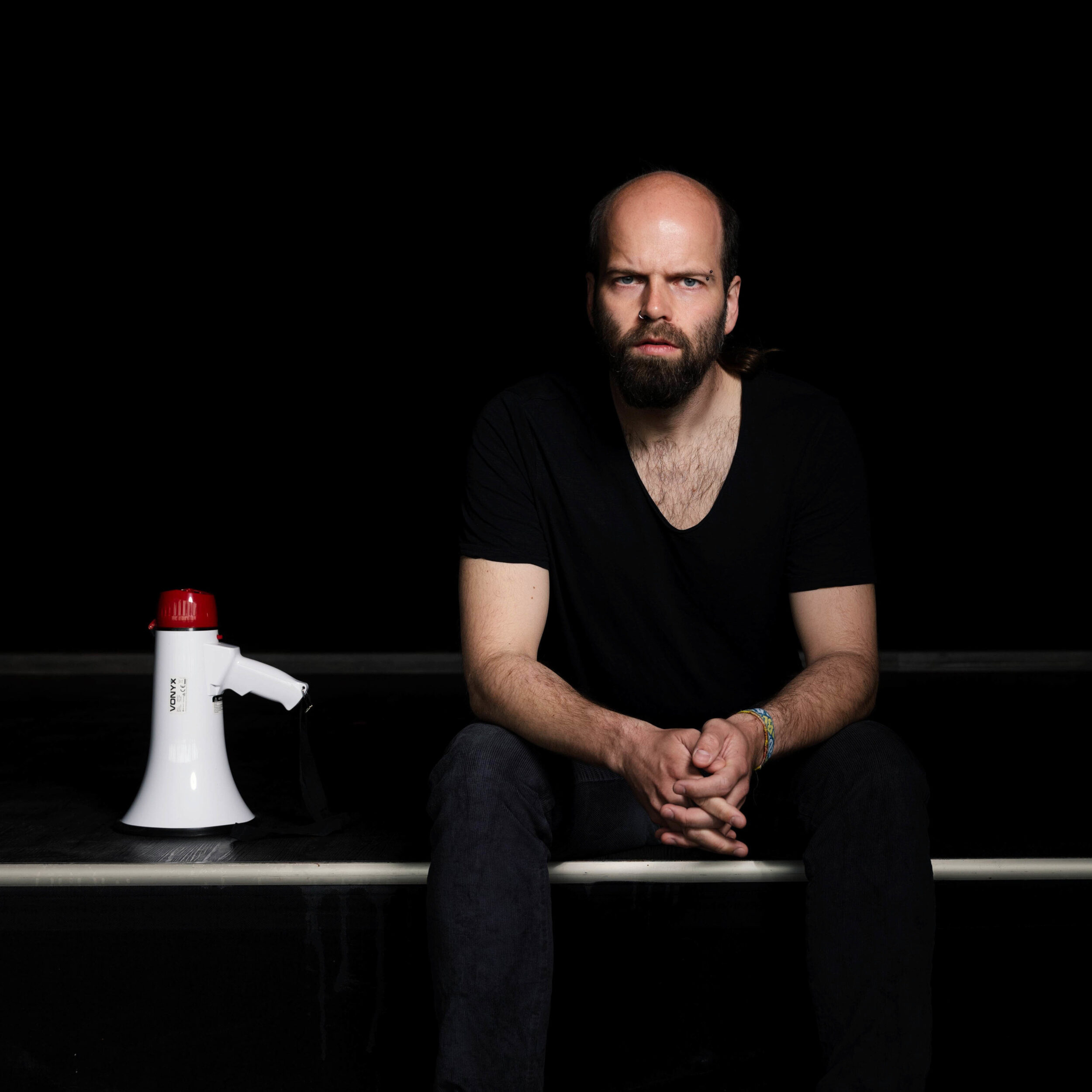 Das Bild zeigt ein Porträt von Pascal Zurek. Er sitzt in schwarzer Kleidung vor einem schwarzen Hintergrund. Links neben ihm liegt ein Megaphon. Er blickt kritisch in die Kamera.