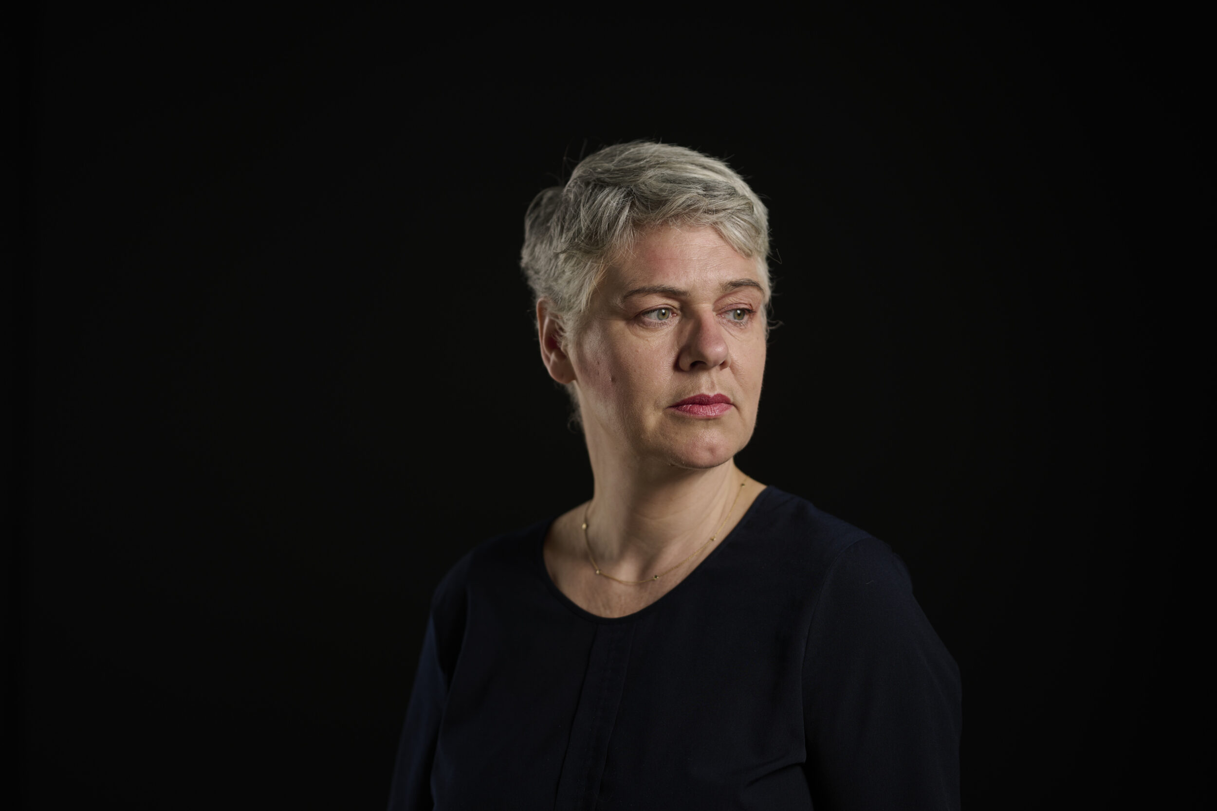 Das Bild zeigt ein Porträt der Mezzosopraistin Truike van der Poel. Sie ist zu sehen mit kurzem, grauem Haar vor einem schwarzen Hintergrund. Sie trägt ein schwarzes Oberteil und blickt zur Seite.