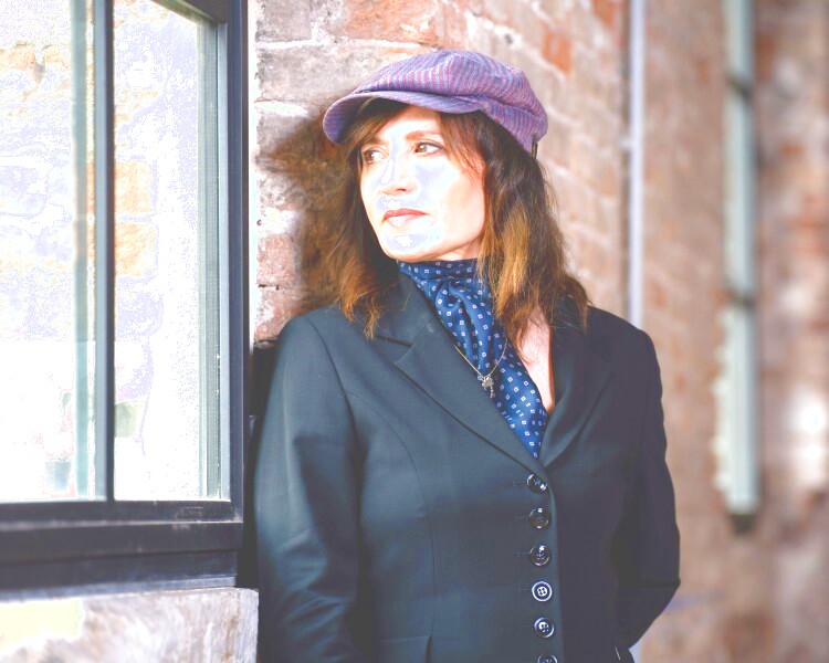 Zu sehen ist ein Portrait der Kompnistin Lucia Ronchetti. Sie trägt eine lilane Schildmütze und ein schwarzes Oberteil mit einem dunkelblauen Schal. Sie lehnt an einer Backsteinwand und blickt in das Fenser neben ihr hinein.