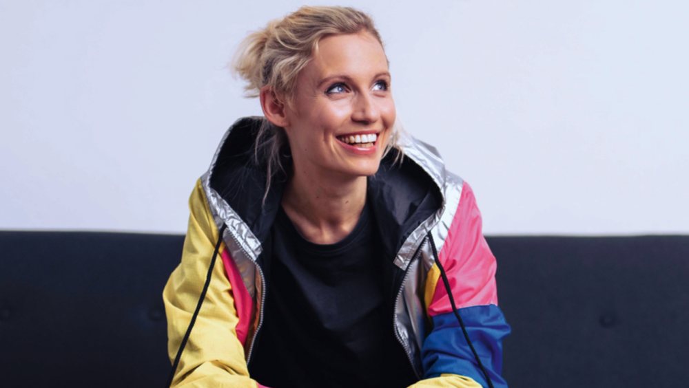 Das Bild zeigt Friederike Scheunchen. Sie trägt einen blonden Zopf und blickt lachend zur Seite. Sie trägt eine bunte Sportjacke in verschiedenen Farben - gelb, pink, blau und silber - über einem schwarzen T-Shirt. Der Hintergrund ist schlicht in Weiß und Grau gehalten.