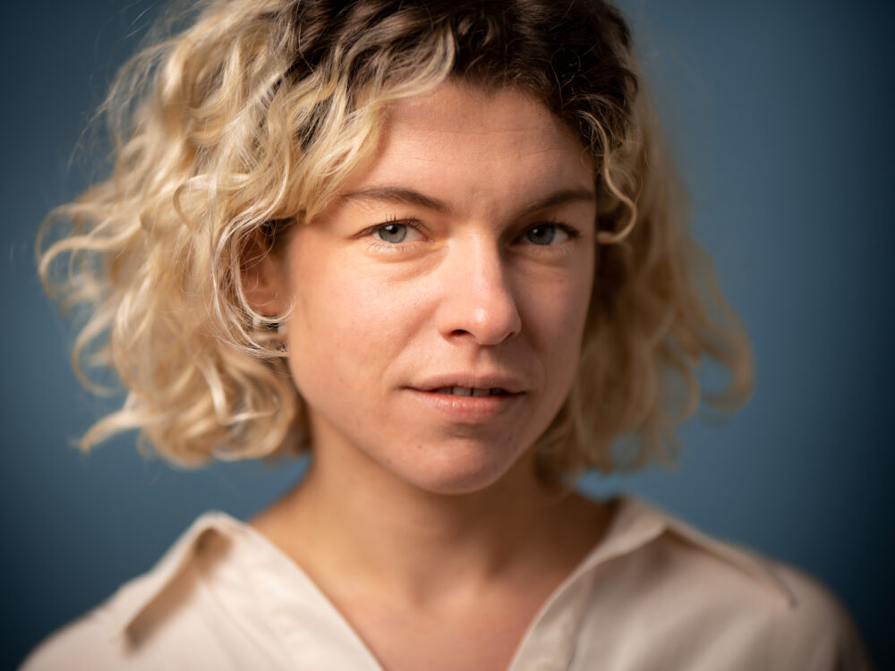 Das Bild zeigt die Autorin Marie Gamillscheg mit kurzen, blonden, lockigen Haaren und blauen Augen. Sie trägt ein helles, beiges Oberteil und blickt direkt in die Kamera. Der Hintergrund ist blau.