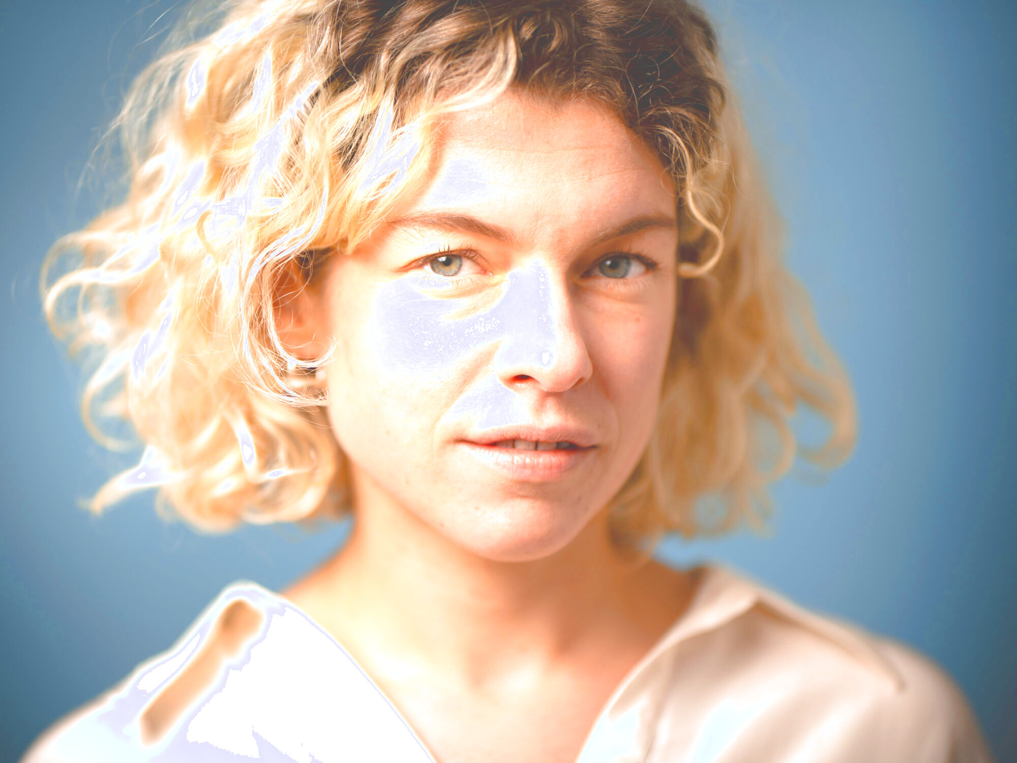 Das Bild zeigt die Autorin Marie Gamillscheg mit kurzen, blonden, lockigen Haaren und blauen Augen. Sie trägt ein helles, beiges Oberteil und blickt direkt in die Kamera. Der Hintergrund ist blau.