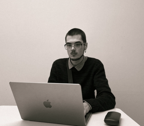 Ilya Viazow mit kurzen Haaren, Brille und Ohrring sitzt an einem Tisch vor einem aufgeklappten Laptop. Er trägt einen dunklen Pullover über einem Hemd und blickt konzentriert auf den Bildschirm. Neben dem Laptop liegt ein Brillenetui. Der Hintergrund ist schlicht und hell.