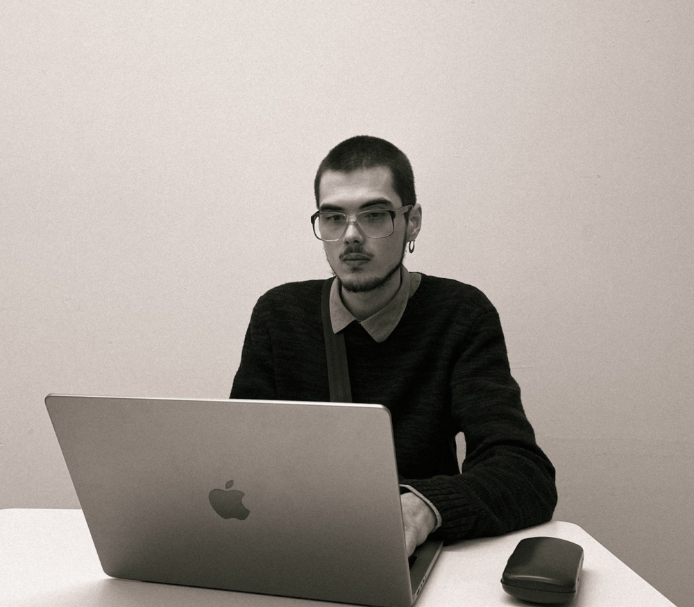 Ilia Viazow mit kurzen Haaren, Brille und Ohrring sitzt an einem Tisch vor einem aufgeklappten Laptop. Er trägt einen dunklen Pullover über einem Hemd und blickt konzentriert auf den Bildschirm. Neben dem Laptop liegt ein Brillenetui. Der Hintergrund ist schlicht und hell.