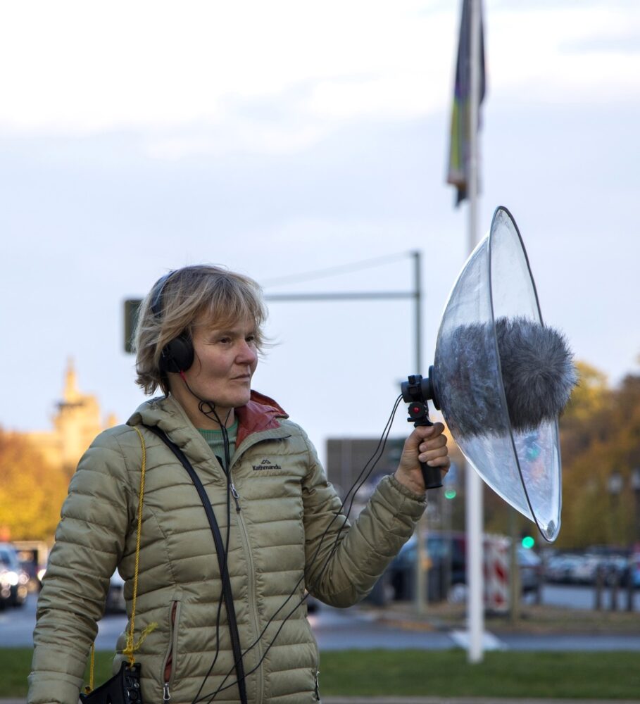 Das Bild zeigt Kirsten Reese. Sie trägt eine beige Jacke, steht im Freien und hält Mikrofon mit Windschutz und Kopfhörern. Im Hintergrund ist eine herbstliche Straßenszene mit gelben Bäumen und Verkehrsampeln zu sehen. Sie scheint Tonaufnahmen zu machen.