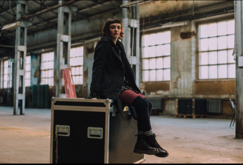 Ludmilla Pogodina sitzt auf einem großen schwarzen Case in einer leeren Industriehalle. Sie hat einen braunen Bob und trägt eine dunkle Jacke, ein gemustertes Kleid, rote und dunkelblaue Strumpfhosen sowie braune Stiefel mit karierten Socken. Die Halle ist lichtdurchflutet, mit hohen Fenstern, abblätternder Wandfarbe und sichtbaren Stahlträgern.