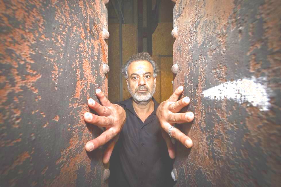 Das Bild zeigt Samir Odeh-Tamimi mit grau meliertem Haar und Bart. Er steht zwischen zwei großen, verwitterten Metallpfeilern. Er trägt ein schwarzes Hemd und streckt beide Hände mit ausgebreiteten Fingern durch die Pfeiler in Richtung der Kamera aus.