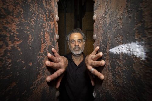 Das Bild zeigt Samir Odeh-Tamimi mit grau meliertem Haar und Bart. Er steht zwischen zwei großen, verwitterten Metallpfeilern. Er trägt ein schwarzes Hemd und streckt beide Hände mit ausgebreiteten Fingern durch die Pfeiler in Richtung der Kamera aus.