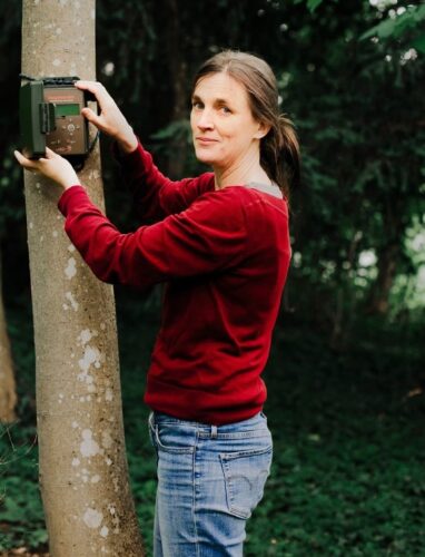 Das Bild zeigt Dr. Sandra Müller in rotem Pullover und Jeans befestigt ein Audioaufnahmegerät an einem Baumstamm. Sie arbeitet in einem Waldgebiet und scheint Naturgeräusche aufzunehmen.