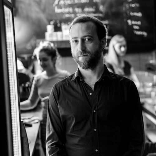 Zu sehen ist ein schwarzweißes Portrait des Komponisten Sharif Sehnaoui. Er trägt ein schwarzes Hemd und blickt freundlich in die Kamera. Im Hintergrund sind verschwommen zwei Frauen zu sehen.