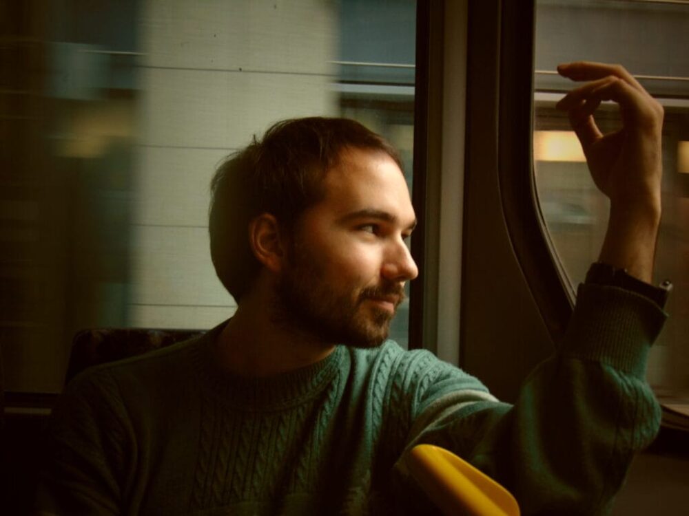 Das Bild zeigt Mert Morali mit dunklen Haaren und Bart in einem türkisfarbenen Strickpullover. Er sitzt an einem Straßenbahnfenster und blickt nachdenklich zur Seite, während er eine Hand an den Kopf hält.